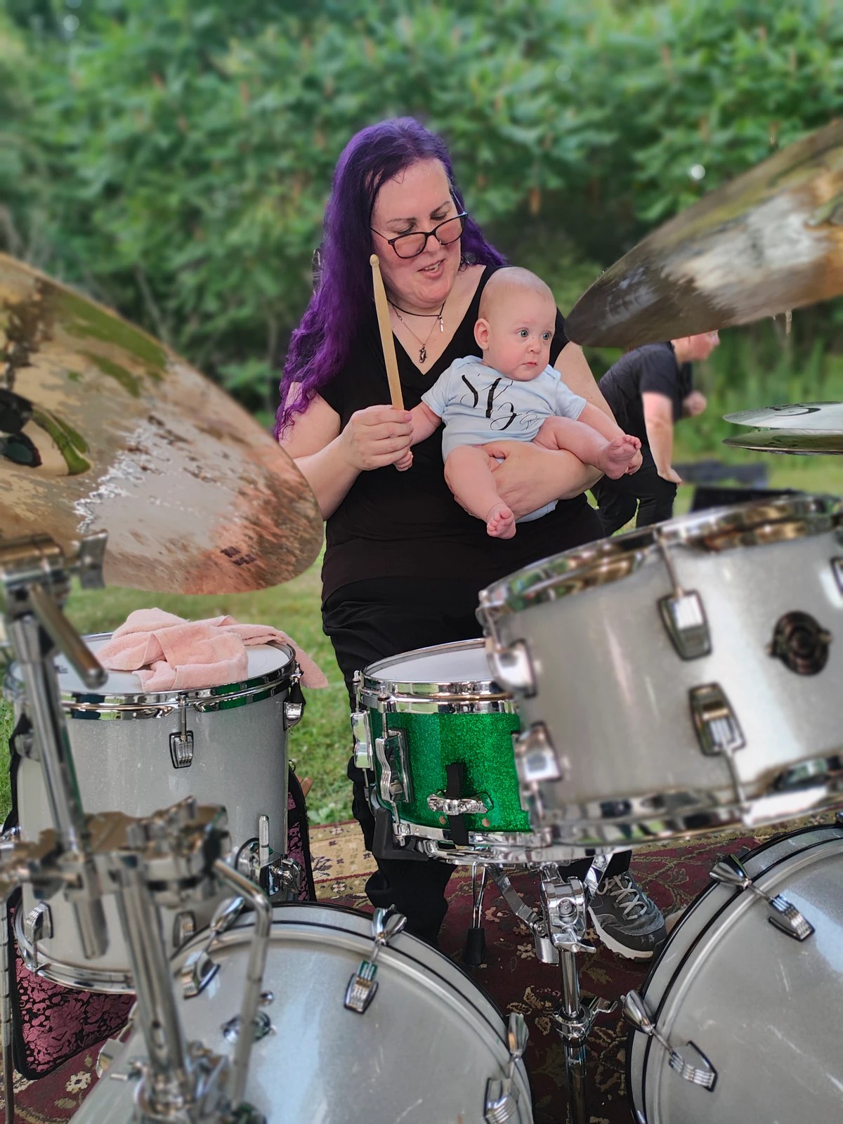 Penny Larson playing drums outdoors while holding her baby