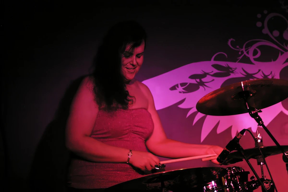 Penny Larson playing drums at a Boston club under purple stage lights